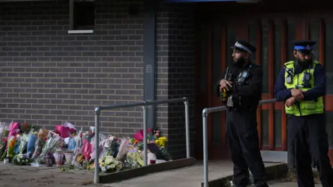 PA Media Two male police officers stand outside the Leabank flats entrance, near to the floral tributes dzzdyzeqzydzzyzdzzyerzdyzezxytqxzyxeuuinv eiqrqikikzprw