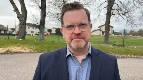 Picture of Calum MacPherson with his head and shoulders in the frame. He has brown hair, a grey bear and glasses and is wearing a dark blue blazer with a lighter blue shirt underneath.