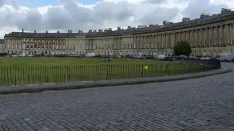 The Royal Crescent in Bath as seen from the street. It is a grand sweeping row of terraced houses with a park and railings in front