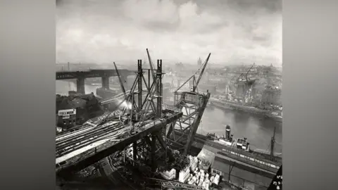 A black and white photograph showing the building on the Tyne Bridge from the Gateshead side. Construction is taking place on either side of the river, but the arch going over the water is just beginning to take shape. There are factories and buildings surrounding the structure, with the High Level Bridge and Swing Bridge visible behind it to the left. There are multiple cranes working on the bridge. The sky is covered in white clouds.