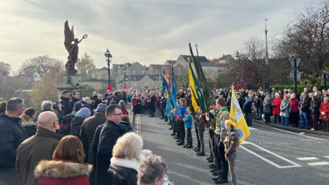 A large outdoor Remembrance Day parade with crowds lining both sides of a road. In the center, a group of uniformed individuals stand in formation holding colorful flags, including blue, green, and yellow banners. Behind them, more participants march along the street.