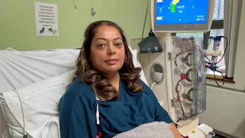 A 45-year-old woman is sitting in a hospital bed next to a dialysis machine. She is wearing a blue jumper and has long dark hair. She is sitting under a blanket.