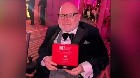 Jason Wilsher-Mills holding his award and smiling at the camera, wearing a bow tie and a suit