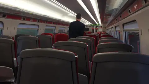 Inside a train carriage with the rear of a train conductor walking along an aisle passing empty seats. The rear of the seats are grey and the cushions are red.