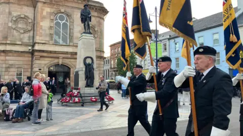 Three men, dressed in navy suits, walking with flags. People are gathered at a statue in the background. 