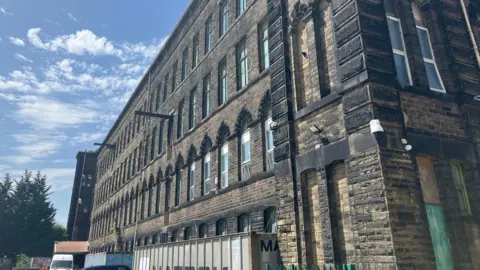Grace Wood/BBC A large mill building with five floors. The stone is dark and smoke stained. There are shipping containers outside