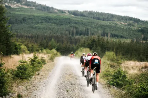 Red On Sports Cyclists race along a dusty track with the tree-covered Galloway hills in the background