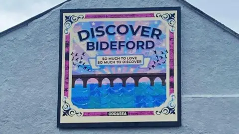 The 2m by 2m artwork sits at the gable end of Country Cottage Bakery. It is a white background with a bridge, water and DISCOVER BIDEFORD - written above it in all capital letters. There are purple and blue stripes in the design.