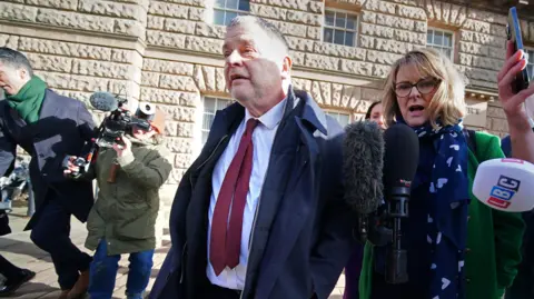 PA Media Mike Amesbury, wearing a navy blue coat over a white shirt and burgundy tie, walks away from court flanked by journalists holding microphones and camera operators