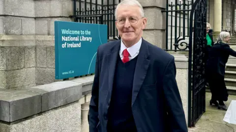 PA Media A white-haired man in a suit walks across the street. 