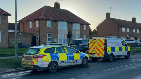 A police car and police forensic van are parked at the side of Hawthorn Drive. Houses can be seen to the left of them with other vehicles parked on a grass verge.