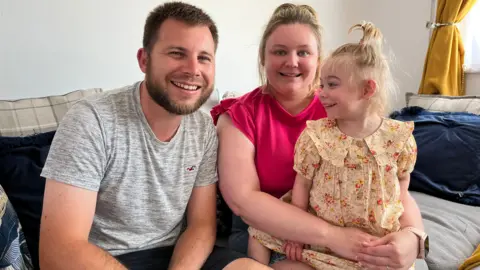 Rich and Jodie with their daughter Amelia. Rich has short dark hair and facial hair. Jodie has blonde hair. Amelia is sat on her lap, looking towards Rich. All of them are smiling.