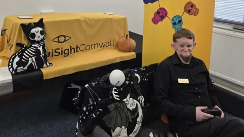 Dominic Hall sitting on a chair. He is wearing a black shirt and black trousers. He is holding a black gaming console. The chair he is sat on has a black blanket with a bat and moon on it. Behind him is a pillow of a cat with a skeleton. There is an iSightCornwall logo behind him and a pumpkin pillow. The walls around him are white and the floor has grey carpet. 