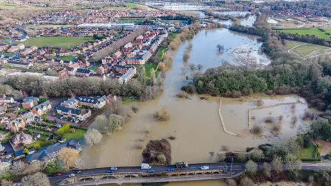 JP/Airwave Drones A drone photo of flooding around Wimborne. Some vehicles are seen driving on a bridge while the green spaces opposite a residential area are flooded.