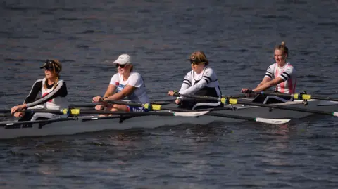 Pacemaker Press Four women are rowing a white boat in a river. Three are wearing caps and two are wearing sunglasses. All three have their hair tied back.