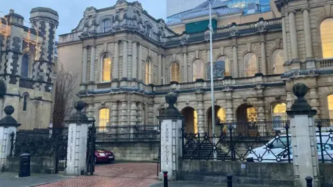 Reading Crown Court: a light brown building with about 15 windows. Lights are shining in most windows. Four pillars stand at the front. Two bear the words "Crown Court". A white van and a red car are parked in front of the building.