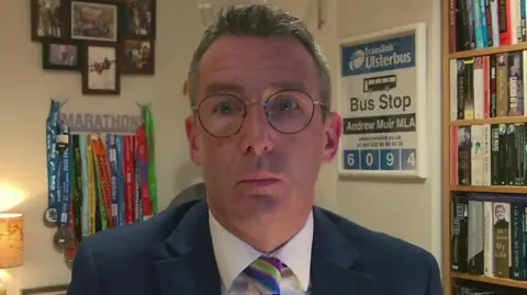 BBC Andrew Muir looking at the camera. He is wearing a navy suit jacket, white shirt, round glasses and multi-colour striped tie. In the background, there is a book shelf filled with books, artwork and medals on the walls and a lamp.