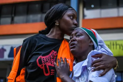 Gerald Anderson / Getty Images A woman embraces and comforts another woman who is crying. Both are at the protest in Nairobi. Behind them are shops.