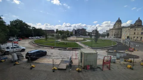 BBC Construction work taking place at Queens Gardens, with lawns and a fountain in the background and construction vehicles and cabins in the foreground.