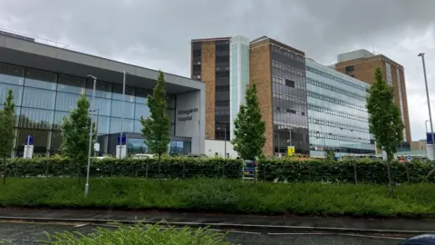 A wide shot of Altnagelvin Hospital, as viewed from a road in front of the building. It consists a multi-storey tower blocks and a large, glass-fronted atrium-style entrance. A number of small trees and a privet-type hedge surround the site. 