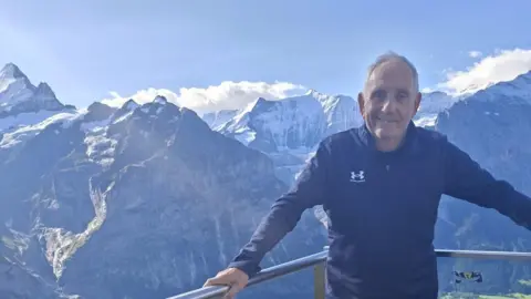 Utopia Travel A man wearing a full-sleeved jacket and trousers, he is smiling at the camera while leaning on a silver railing behind him.
He is stood in front of mountains 