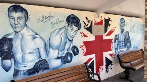 Graffiti art work inside a bus shelter shows three boxers from Chatteris, from left to right, Dave 'Boy' Green, Eric Boon and Jordan Gill. Two wooden benches are in front of the artwork which is done in black and white, with part of a Union flag in the middle.