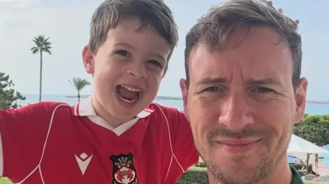 Stephen Rule A man and his young son posing to the camera. The young son is wearing a red Wrexham football shirt. Behind them, we can see the sea and some dotted palm trees.