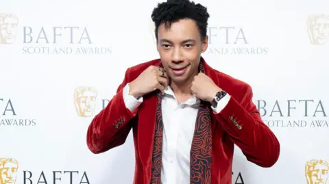 PA Media Kit Young poses at the Bafta Scotland Awards, wearing a red velvet suit and white shirt.