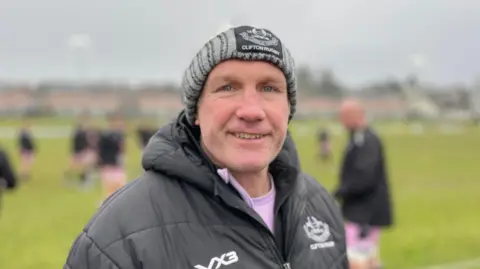 Matt Salter stands on a Rugby pitch wearing a hat and coat with the Clifton Rugby crest on it