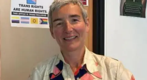 A woman with short grey hair and a cream-coloured blouse with some colourful patches sits in a chair, smiling at the camera. The wall behind her is decorated with pictures and posters, including one which reads: "TRANS RIGHTS ARE HUMAN RIGHTS".