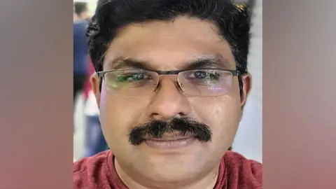 A close-up photograph of Deepak U facing the camera. He has short, dark hair, wears rectangular glasses, and has a thick moustache. His expression is neutral. He is wearing a dark red, short-sleeved T-shirt. The photograph appears to have been taken indoors. Behind him, the background is blurred, with indistinct figures of people and what look like closed shop shutters or metal doors, suggesting a public place