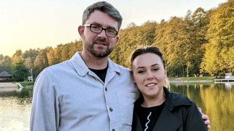 Robbie Anderson stands with his wife in front of a lake. He wears a grey, cord, button-down jacket and glasses and has his arm around his wife who wears a dark grey belted trench coat.