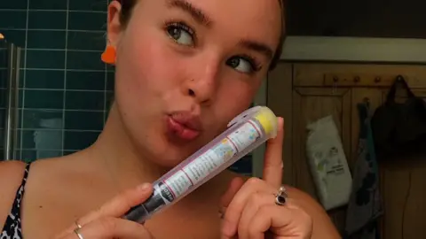 Chloe Dickson Chloe Dickson looks to the left of the camera while holding an EpiPen in a bathroom. It has a black top labelled 'needle' and yellow base, with instructions written on the side.