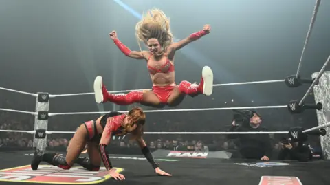 Two women in a wrestling ring. One is down on the floor and the other is jumping over her. There is a camera man behind them outside the ring. 