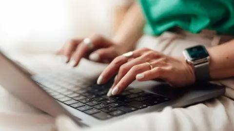 Someone's hands typing on a laptop keyboard. they have white nails and are wearing gold jewellery. They have an apple watch on their rest and are wearing a green top and beige trousers. The laptop is silver.