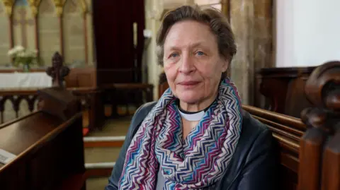 A woman wearing a clerical collar sits in a wooden church pew, looking towards the camera. The Reverend Sue Pulko is wearing a patterned scarf and a dark jacket. Behind her is the interior of a historic church, with carved wooden pews, stone walls and an altar.