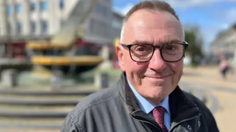 A close up of Tudor Evans. He has short dark hair, is smiling, wearing glasses, a jacket, shirt and red tie.