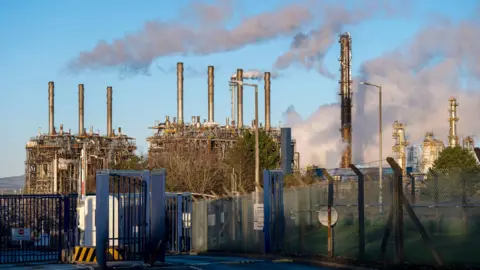 A close-up of the Exxon Mobil site. There are several tall metal cylinders jutting into a blue sky.
