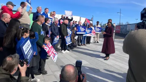 BBC/Luke Walton Kemi Badenoch in a long smart purple coat talks to a crowd of about 50 people holding Conservative Party signs. The Stadium of Light can be see behind her.