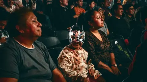 DTU Lucha Libre A child, centre, wearing a Mexican wrestler mask, looking at the camera, with a crowd of people behind him looking to the right