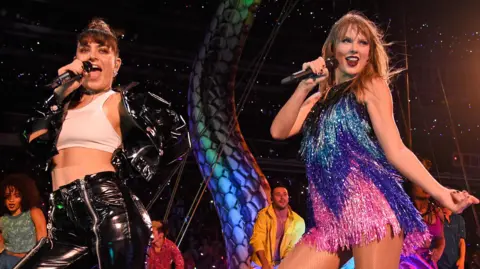Getty Images Charli XCX and Taylor Swift on stage together as part of Taylor's Reputation tour in 2018. Charli wears her dark hair in a top knot and wears a white crop top paired with a patent black jacket and matching trousers. Taylor wears a sparkly mini dress in pink, blue, purple and black. She has long blonde hair and red lipstick. 