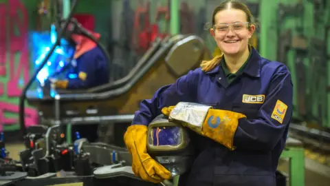 Eleanor Pitt wearing blue JCB overalls, yellow safety gloves and a helmet standing near machinery in a workshop