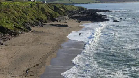 An image of Greenaway Beach, Polzeath. It is a sandy beach. There are people walking along the beach. The sea is blue with white sea foam. To the left of the beach is a grassy hill. 