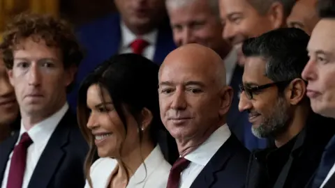 Mark Zuckerberg and Jeff Bezos wearing similar navy suits, with white dress shirts and dark red ties at the second inauguration of Donald Trump. Sundar Pichai and Elon Musk are next to them, both dressed in black.