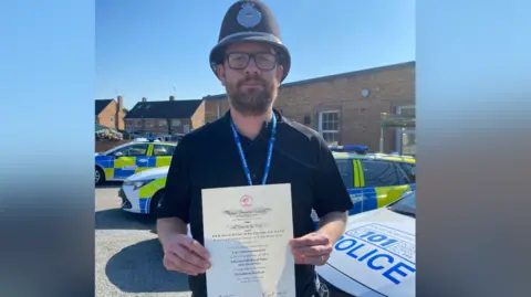A police officer wearing a traditional tall police helmet and a black polo top holds up a white certificate outside with police cars parked behind him.