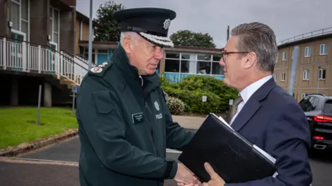 PSNI Jon Boutcher in uniform shakes hands with Keir Starmer who is holding a binder