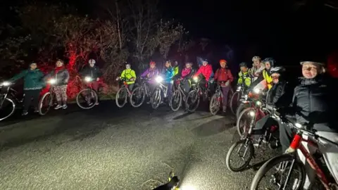 Imogen Calloway, Breeze Network Devon and Cornwall Fifteen cyclists are visible gathered in a semi circle on the road.
They are wearing helmets and their  bike lights are on.