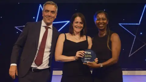 Summerhill Academy Sally Goodridge wearing a black dress and holding her TES award. She is joined on stage by chief judge Jon Severs on her right, wearing a dark suit and red tie. Another woman is on her left, jointly holding the award and smiling.