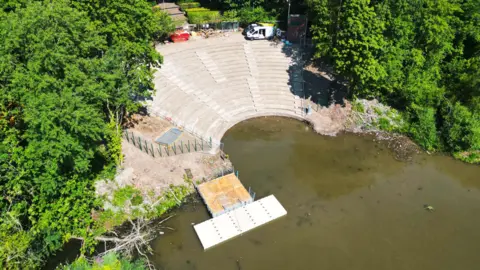 An aerial photo of the amphitheatre show restored circular seating leading to a pool in the park. A pontoon sits on the left with some fencing still in place as work continues. Trees surround the site.