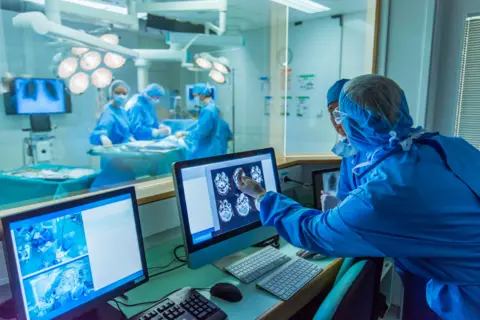 Getty Images Stock Photo dari orang yang melihat gambar otak selama operasi. Di latar belakang adalah teater operasi dengan empat staf dan pasien di atas meja. Ini kabur. Di latar depan ada empat orang yang mengenakan scrub bedah biru yang melihat layar komputer dengan pemindaian otak pada mereka dan satu menunjuk pada satu. 